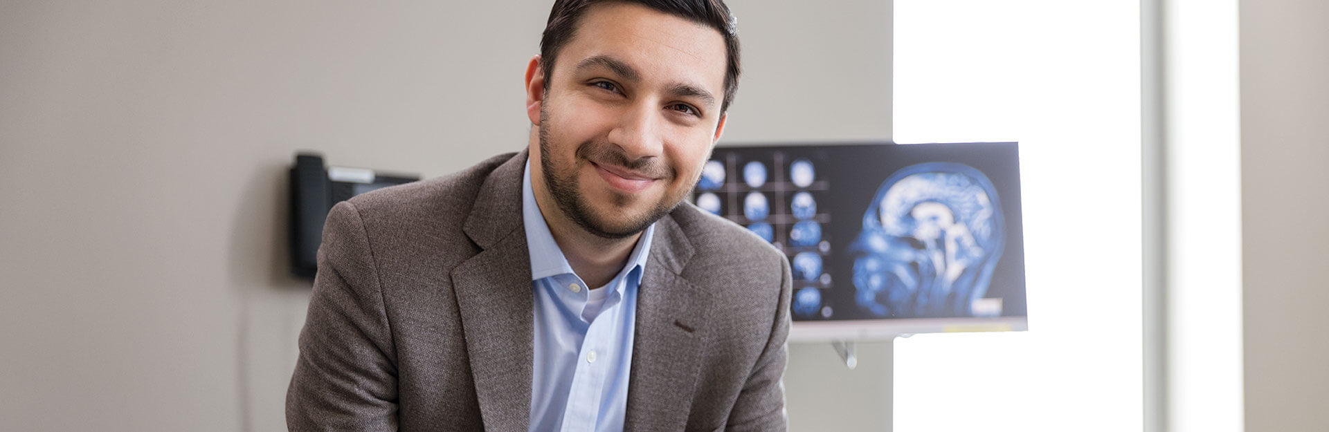 Dr. Ajam sitting in a patient room smiling at the camera with brain scan on computer monitor behind him.